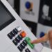Dedo pressionando botão verde "Confirma" em urna eletrônica brasileira, com teclado numérico e painel visíveis