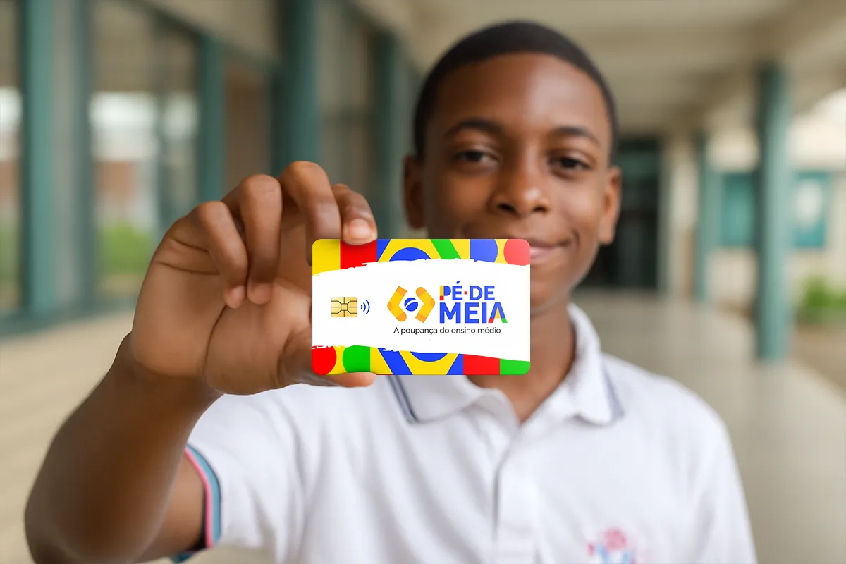 Quer usar o Pé-de-Meia? Veja como conferir se sua conta está liberada 2 Jovem estudante negro sorridente com camisa polo branca segura cartão Pé-de-Meia colorido em ambiente escolar