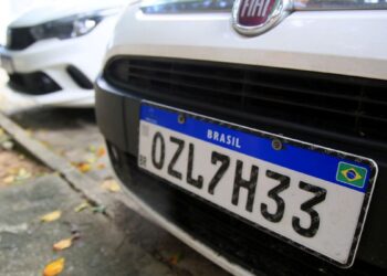 Detalhe em close da parte frontal de um carro branco exibindo uma placa no padrão Mercosul com a combinação OZL7H33 e a bandeira do Brasil no canto inferior direito.