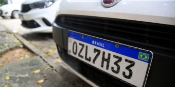 Detalhe em close da parte frontal de um carro branco exibindo uma placa no padrão Mercosul com a combinação OZL7H33 e a bandeira do Brasil no canto inferior direito.