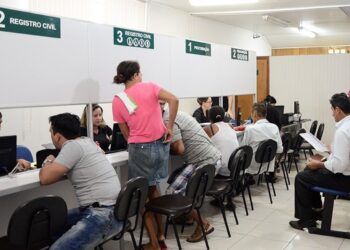 Cidadãos aguardam atendimento em guichês de registro civil com placas verdes numeradas e funcionários em computadores