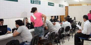 Cidadãos aguardam atendimento em guichês de registro civil com placas verdes numeradas e funcionários em computadores