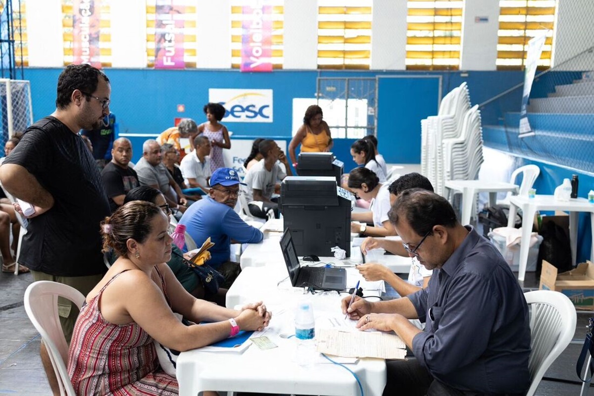 Semana do registro civil: cartórios de todo o Brasil fazem mutirão de 13 a 17 de abril 2 Atendentes auxiliam mulher com documentação em mesa de mutirão de registro civil em ginásio esportivo